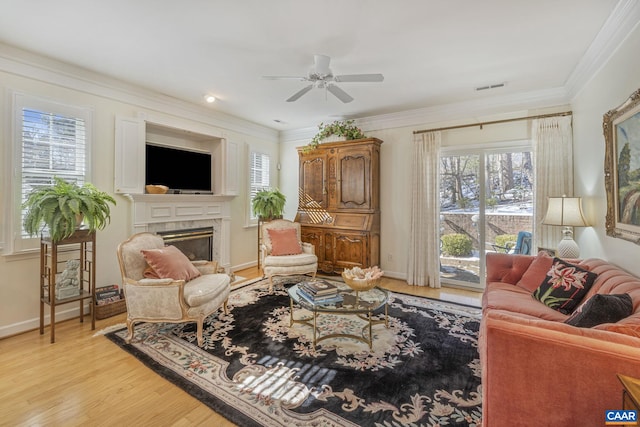 living room with a ceiling fan, visible vents, light wood finished floors, a glass covered fireplace, and crown molding