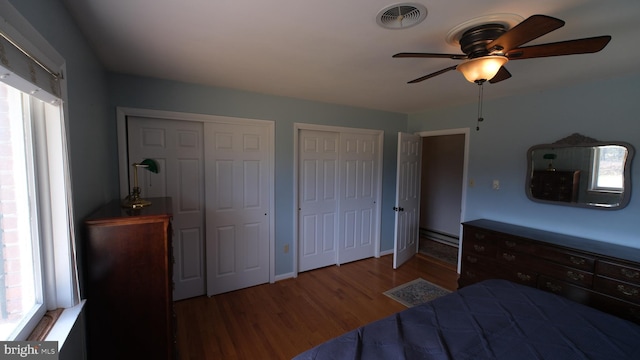 bedroom featuring two closets, baseboard heating, ceiling fan, visible vents, and wood finished floors