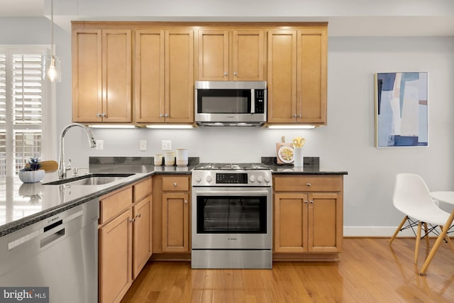 kitchen featuring light wood finished floors, stainless steel appliances, a sink, dark stone counters, and hanging light fixtures
