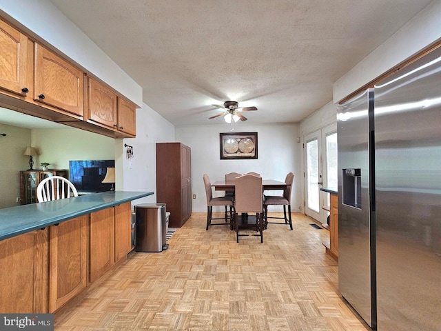 kitchen with ceiling fan, stainless steel refrigerator with ice dispenser, a textured ceiling, dark countertops, and brown cabinets