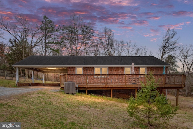 back of property at dusk featuring central AC unit, gravel driveway, a carport, a deck, and a lawn