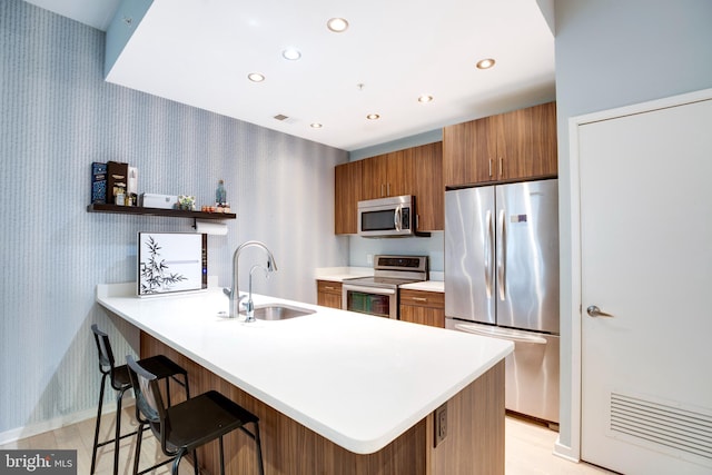 kitchen featuring brown cabinetry, wallpapered walls, a peninsula, a sink, and appliances with stainless steel finishes