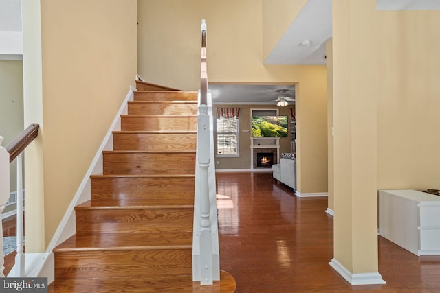 staircase featuring a warm lit fireplace, baseboards, ceiling fan, and wood finished floors