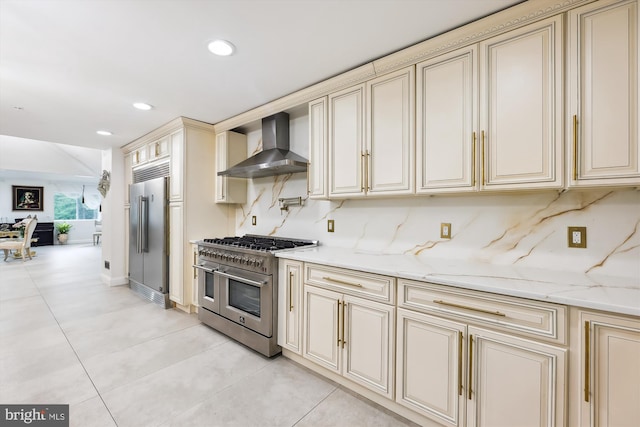 kitchen with premium appliances, cream cabinetry, decorative backsplash, and wall chimney range hood