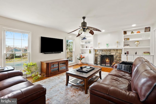 living room with built in shelves, baseboards, ceiling fan, a stone fireplace, and wood finished floors