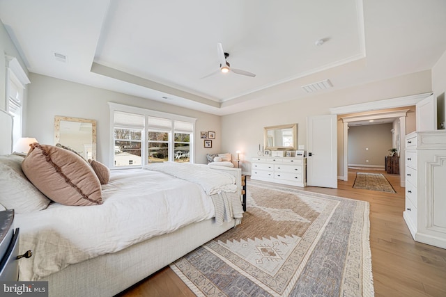 bedroom with a tray ceiling, visible vents, light wood-style flooring, and a ceiling fan