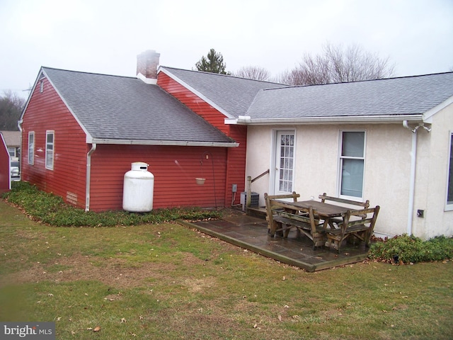 rear view of property featuring a shingled roof, a lawn, stucco siding, a chimney, and a patio area