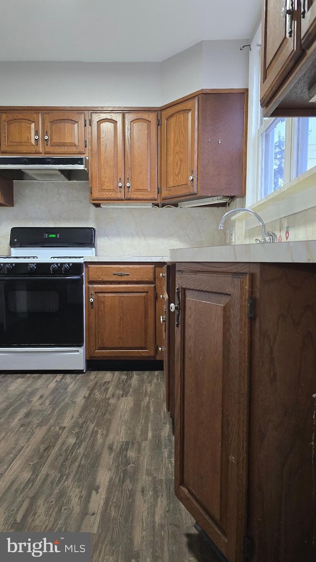 kitchen with brown cabinets, dark wood-style flooring, under cabinet range hood, range with gas cooktop, and tasteful backsplash