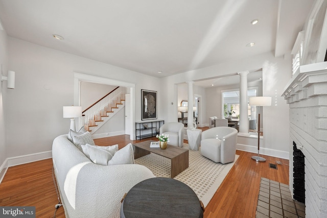 living room featuring a brick fireplace, baseboards, stairs, decorative columns, and wood finished floors