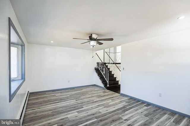 empty room with a baseboard heating unit, baseboards, ceiling fan, stairway, and wood finished floors