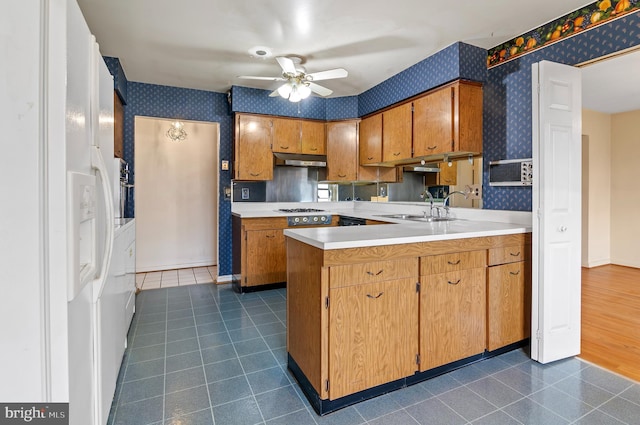 kitchen with a ceiling fan, wallpapered walls, a peninsula, a sink, and under cabinet range hood