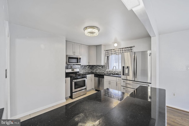 kitchen with backsplash, baseboards, dark stone counters, appliances with stainless steel finishes, and a sink