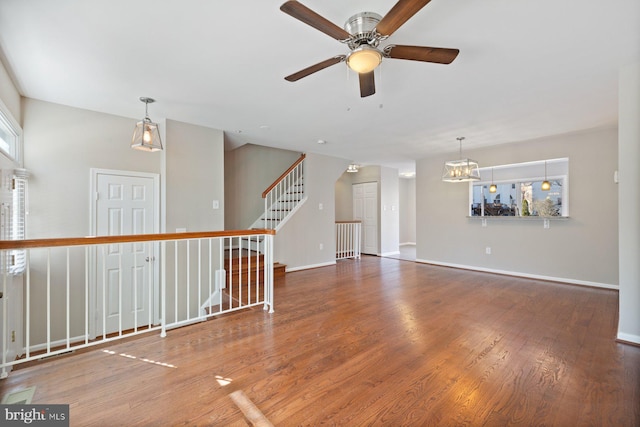 unfurnished living room with wood finished floors, stairway, ceiling fan with notable chandelier, and baseboards