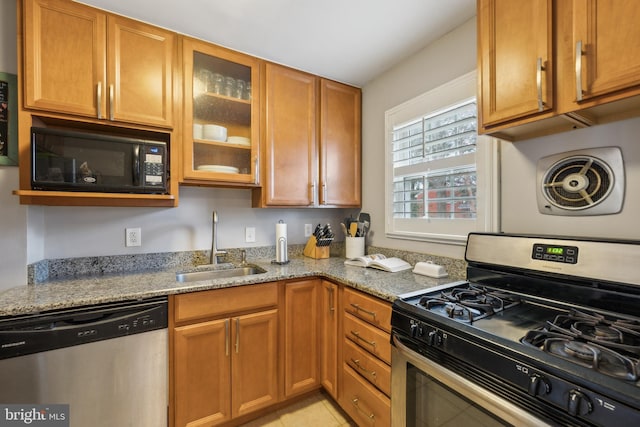 kitchen with a sink, light stone counters, stainless steel appliances, brown cabinetry, and glass insert cabinets