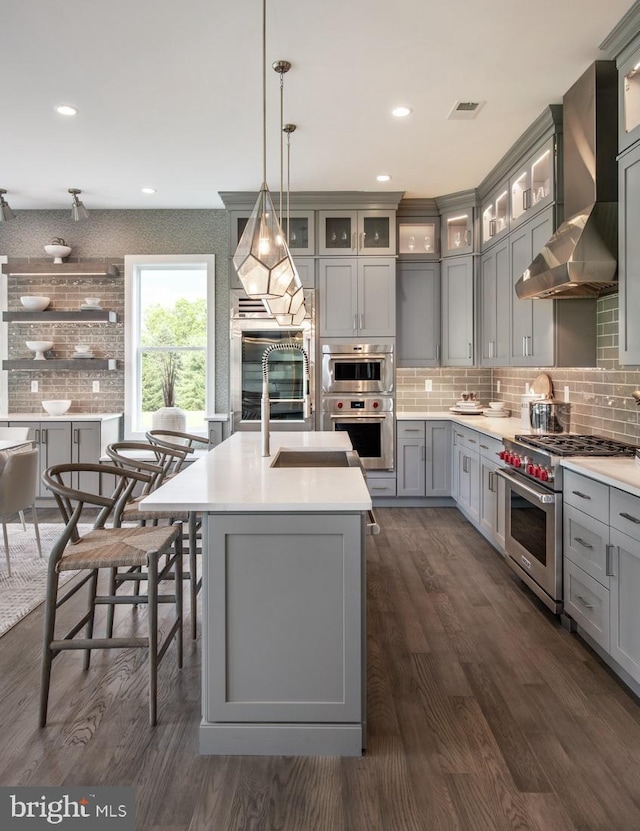 kitchen with backsplash, high end stove, wall chimney range hood, light countertops, and gray cabinets