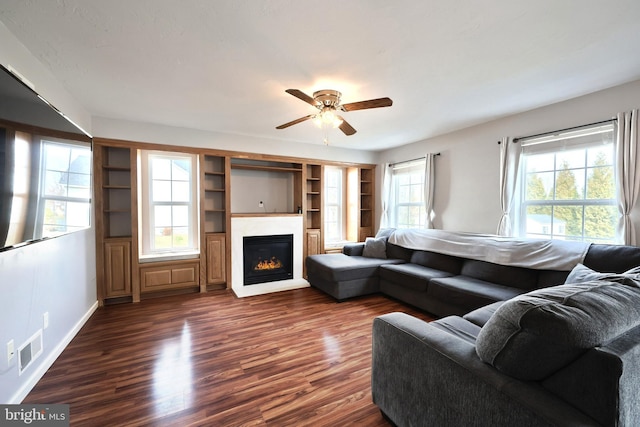 living area featuring visible vents, a ceiling fan, a glass covered fireplace, baseboards, and dark wood-style flooring