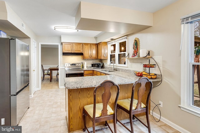 kitchen with under cabinet range hood, light stone counters, decorative backsplash, a peninsula, and stainless steel appliances