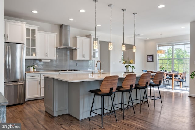 kitchen featuring tasteful backsplash, a sink, stainless steel refrigerator, wall chimney exhaust hood, and a center island with sink
