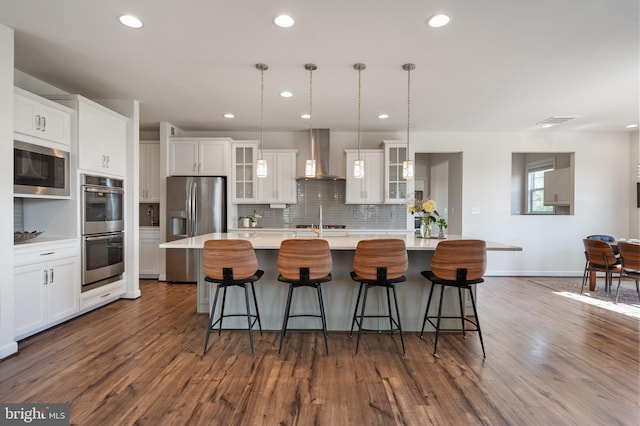 kitchen featuring stainless steel appliances, tasteful backsplash, wall chimney range hood, light countertops, and an island with sink