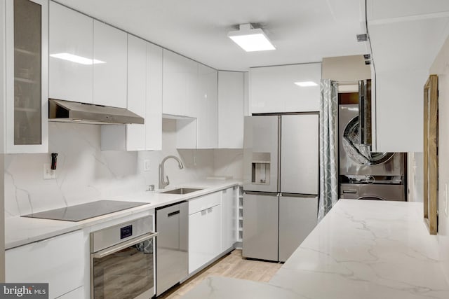 kitchen featuring a sink, white cabinets, under cabinet range hood, appliances with stainless steel finishes, and stacked washer / dryer