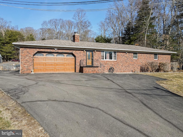 ranch-style home with aphalt driveway, an attached garage, a shingled roof, brick siding, and a chimney