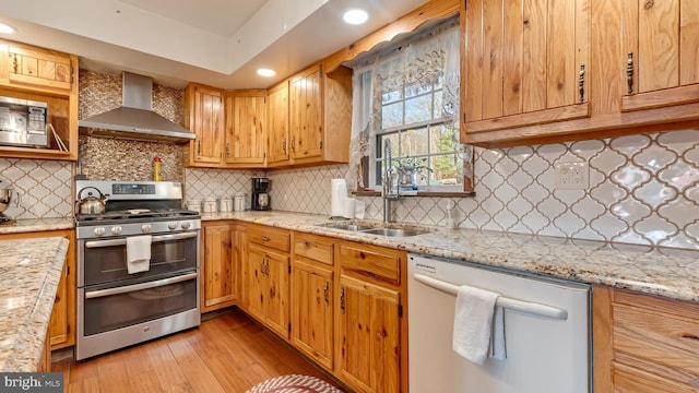 kitchen featuring light wood finished floors, wall chimney range hood, light stone countertops, stainless steel appliances, and a sink