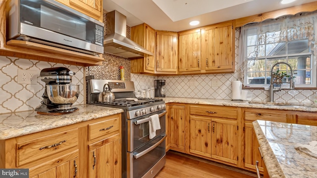 kitchen with light wood-type flooring, a sink, appliances with stainless steel finishes, wall chimney range hood, and decorative backsplash