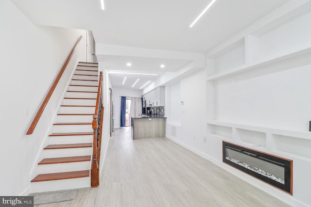 unfurnished living room featuring recessed lighting, light wood-style floors, a glass covered fireplace, and stairs