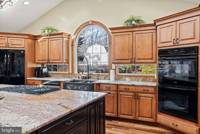 kitchen with light wood-type flooring, vaulted ceiling, light stone counters, black appliances, and a sink