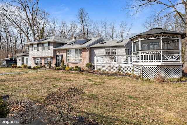 view of front of house with roof mounted solar panels, roof with shingles, a front yard, a wooden deck, and a chimney