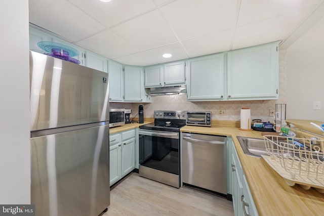 kitchen featuring a toaster, stainless steel appliances, under cabinet range hood, light wood-type flooring, and backsplash