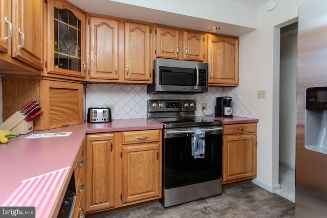 kitchen with baseboards, stainless steel appliances, glass insert cabinets, tasteful backsplash, and brown cabinets