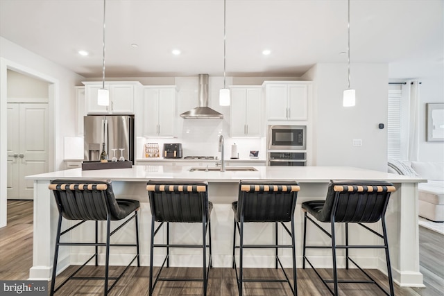 kitchen with wall chimney exhaust hood, stainless steel appliances, a large island with sink, a sink, and wood finished floors