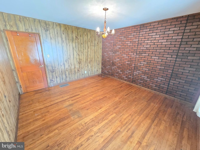 unfurnished room with a notable chandelier, visible vents, brick wall, and wood finished floors