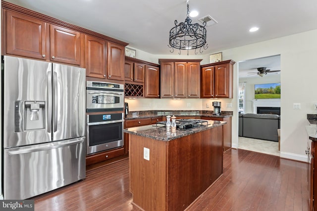 kitchen with dark stone counters, appliances with stainless steel finishes, ceiling fan with notable chandelier, dark wood-style flooring, and a lit fireplace
