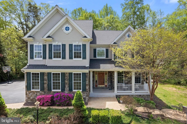 traditional home featuring covered porch, roof with shingles, stucco siding, and stone siding