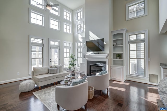 living area featuring baseboards, dark wood finished floors, a high ceiling, ceiling fan, and a glass covered fireplace