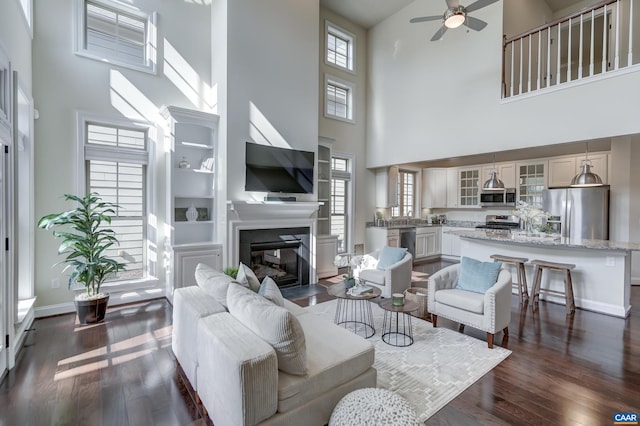 living room featuring ceiling fan, baseboards, a glass covered fireplace, and dark wood-style floors