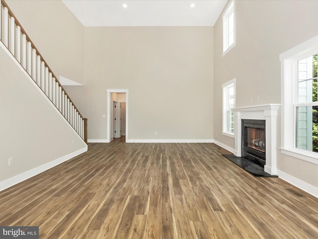 unfurnished living room with stairway, a fireplace with raised hearth, baseboards, a towering ceiling, and wood finished floors