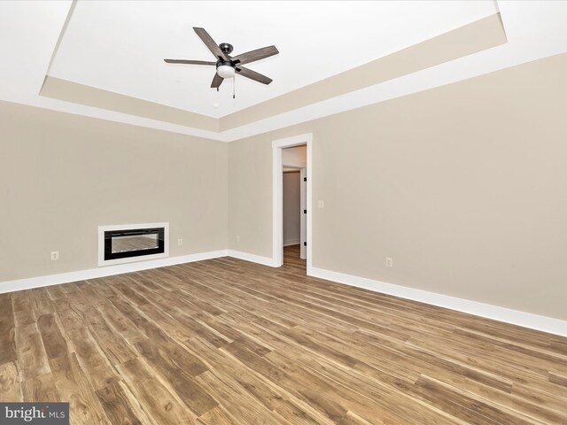 unfurnished living room with a raised ceiling, a glass covered fireplace, a ceiling fan, baseboards, and wood finished floors