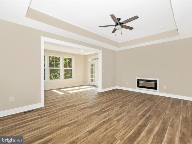 unfurnished living room featuring ceiling fan, a raised ceiling, visible vents, baseboards, and wood finished floors