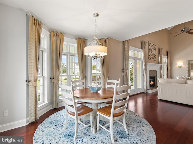dining space featuring baseboards, dark wood finished floors, lofted ceiling, ceiling fan with notable chandelier, and a warm lit fireplace