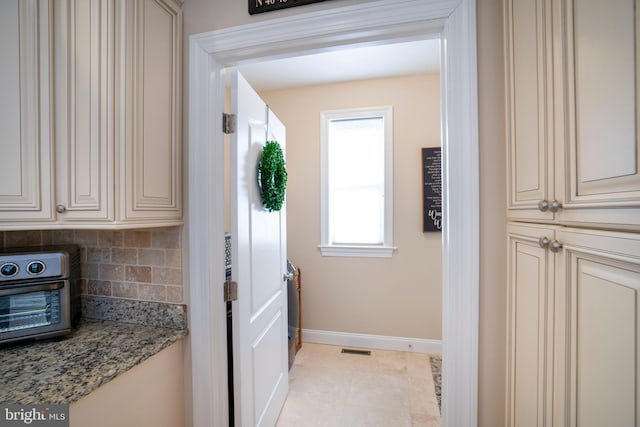 kitchen featuring baseboards, a toaster, decorative backsplash, cream cabinetry, and stone countertops