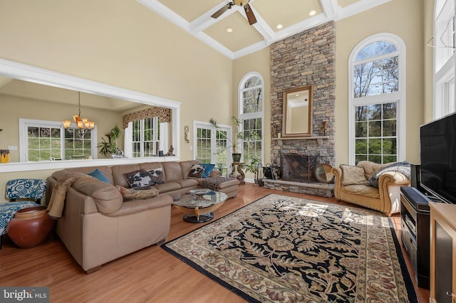 living room featuring light wood finished floors, ceiling fan with notable chandelier, beamed ceiling, a high ceiling, and a stone fireplace
