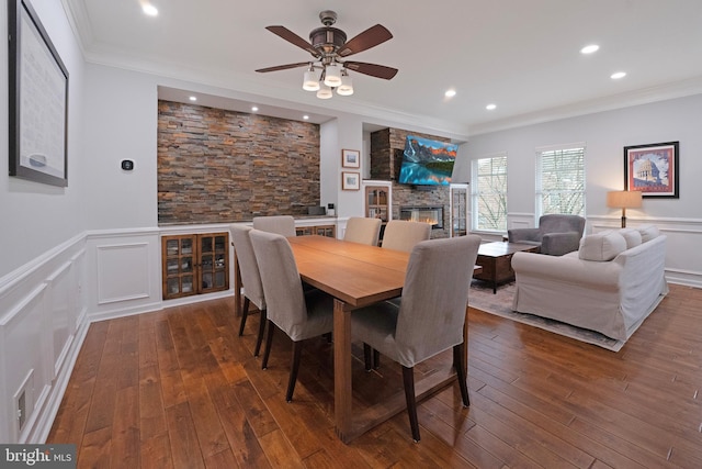 dining area with a decorative wall, ornamental molding, a ceiling fan, a fireplace, and dark wood finished floors