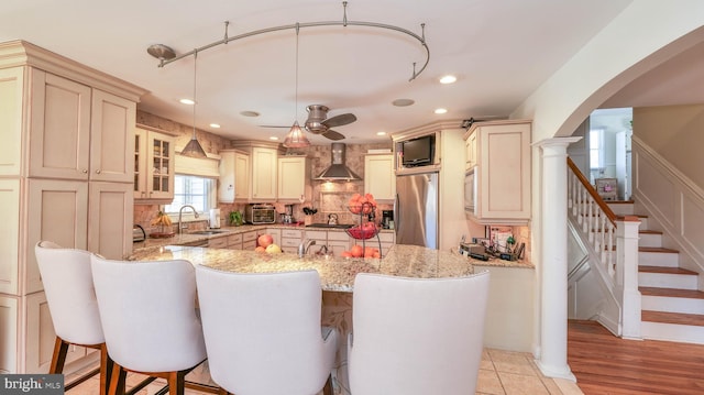 kitchen with a breakfast bar area, cream cabinets, stainless steel appliances, wall chimney exhaust hood, and a sink