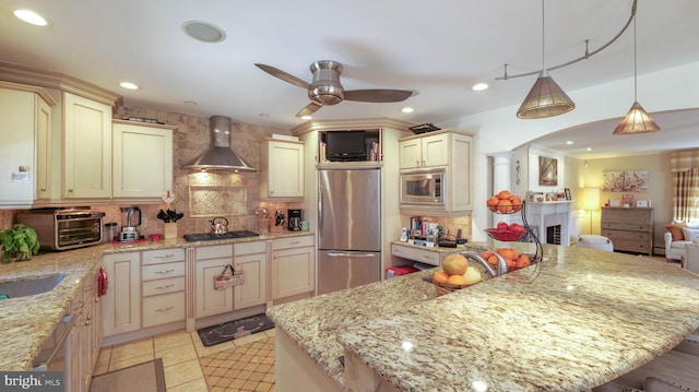 kitchen featuring ceiling fan, decorative backsplash, appliances with stainless steel finishes, cream cabinets, and wall chimney range hood
