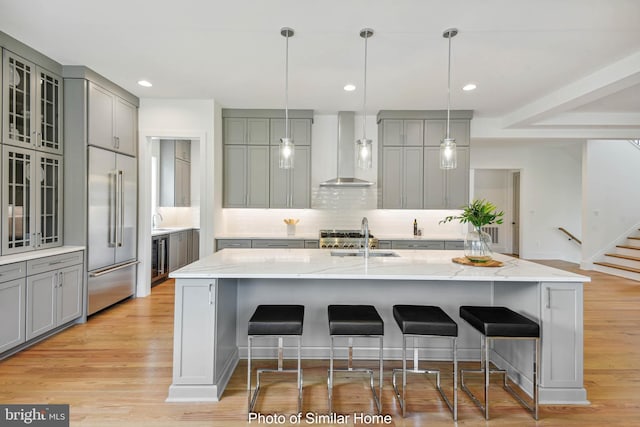 kitchen with light stone counters, a sink, wall chimney exhaust hood, gray cabinetry, and stainless steel built in refrigerator
