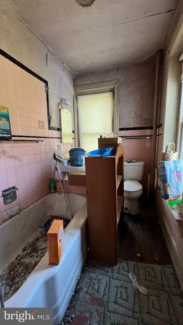 bathroom with tile walls, a textured ceiling, toilet, wainscoting, and a tub to relax in
