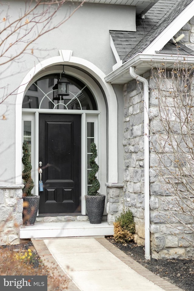 property entrance with stone siding, roof with shingles, and stucco siding
